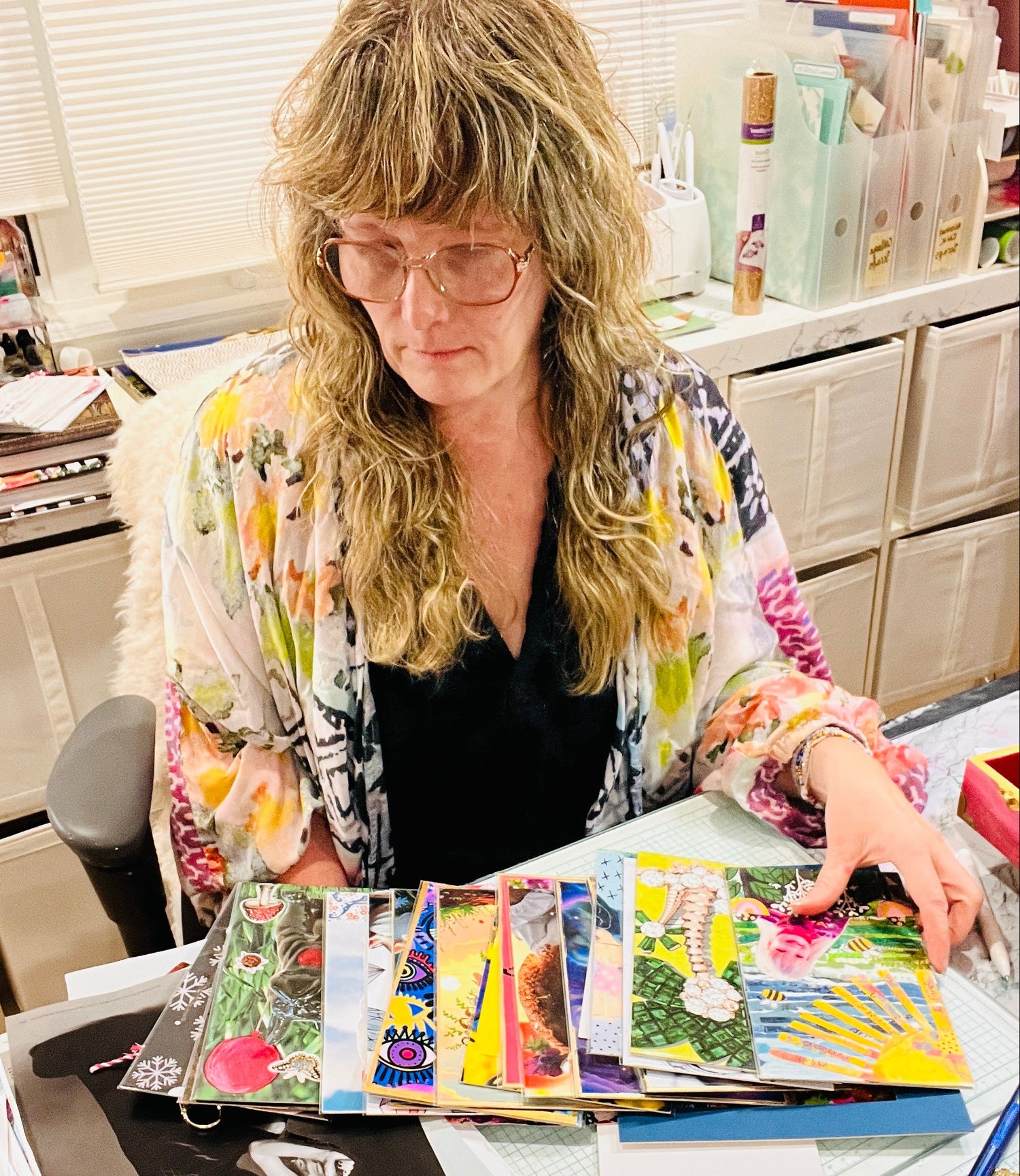 A woman with blonde hair and glasses sits at a desk surrounded by colorful 
SoulCollage® cards, papers, and art supplies in a bright craft or studio room.